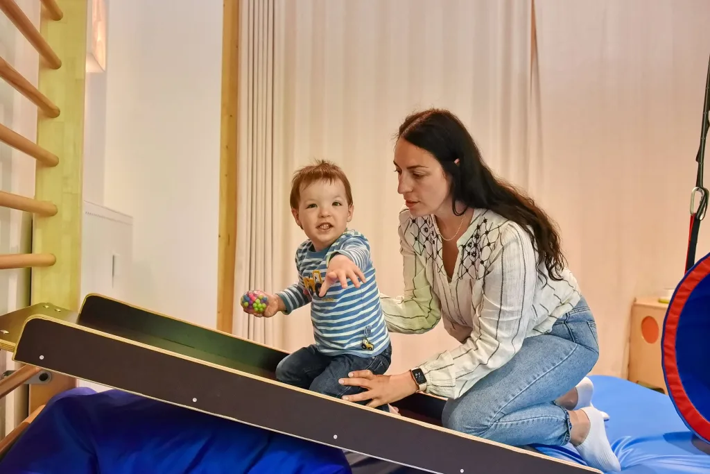 Fotoaufnahme eines Kindes zusammen mit einer Betreuerin im Innenbereich auf einer Wippe