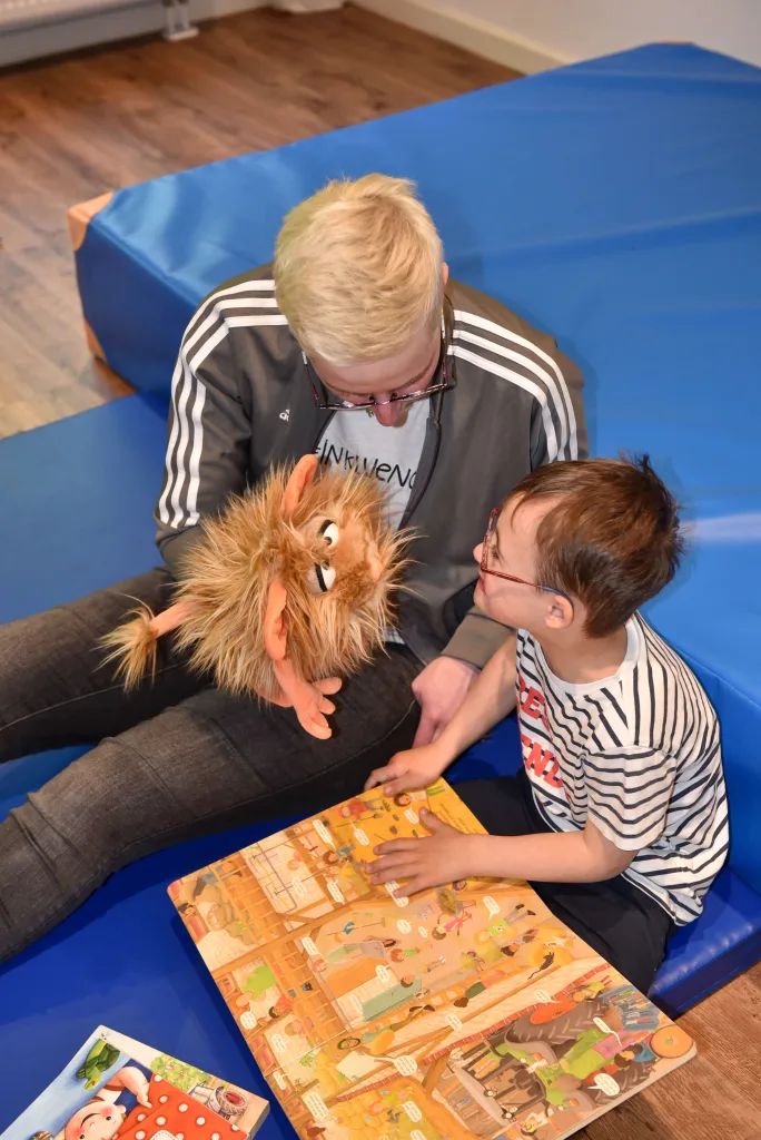 Fotoaufnahme einer Betreuerin mit einer Handpuppe und eines Kindes mit einem Buch auf dem Schoß