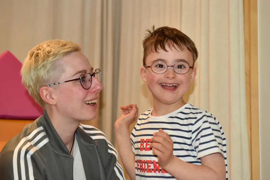 Fotoaufnahme eines glücklichen Kindes, das in die Kamera strahlt; die Betreuerin schaut das Kind fröhlich an